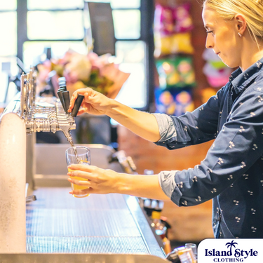 Female bartender wearing Palm & Pawn Hotel custom hospitality uniform pouring beer at bar. Branded long sleeve shirt for food service staff.
