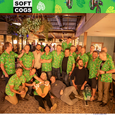 Group of men wearing Soft Cogs Sports Uniforms custom Hawaiian shirts in green with cartoon prints, gathered at a social event indoors.