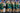 Group of people in green and blue uniforms with flags, likely at a sports event.