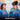 Two women smiling and looking at each other, wearing matching blue tropical print short-sleeve collared shirts with pink, yellow, and green floral motifs. A blurred GBA office background is visible behind them, with the word 'Anniversary' overlaid at the bottom.