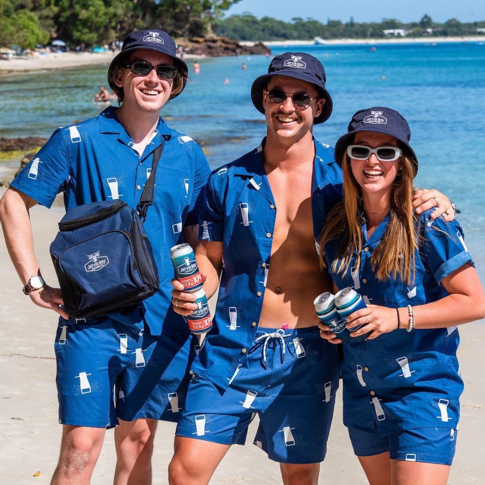 Three people in matching blue outfits with sunglasses and drinks by a beach.