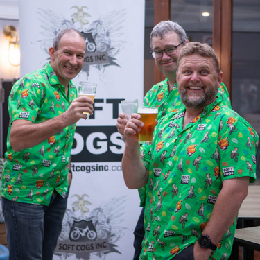 Three men smiling and holding beers while wearing Soft Cogs Sports Uniforms custom Hawaiian shirts with vibrant green cycling-themed design.