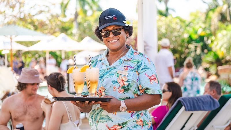 Waitress in a tropical setting holding drinks and a tray with people in the background
